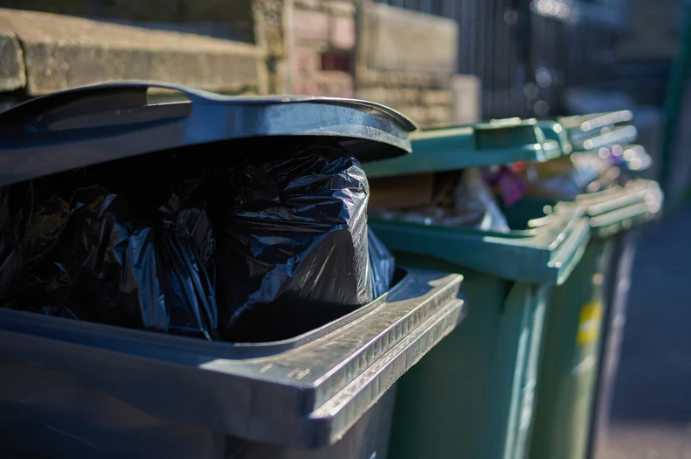 Three garbage bins lined up against a wall: the first black bin has black bags inside with the lid open; the second and third green bins contain loose, unbagged garbage